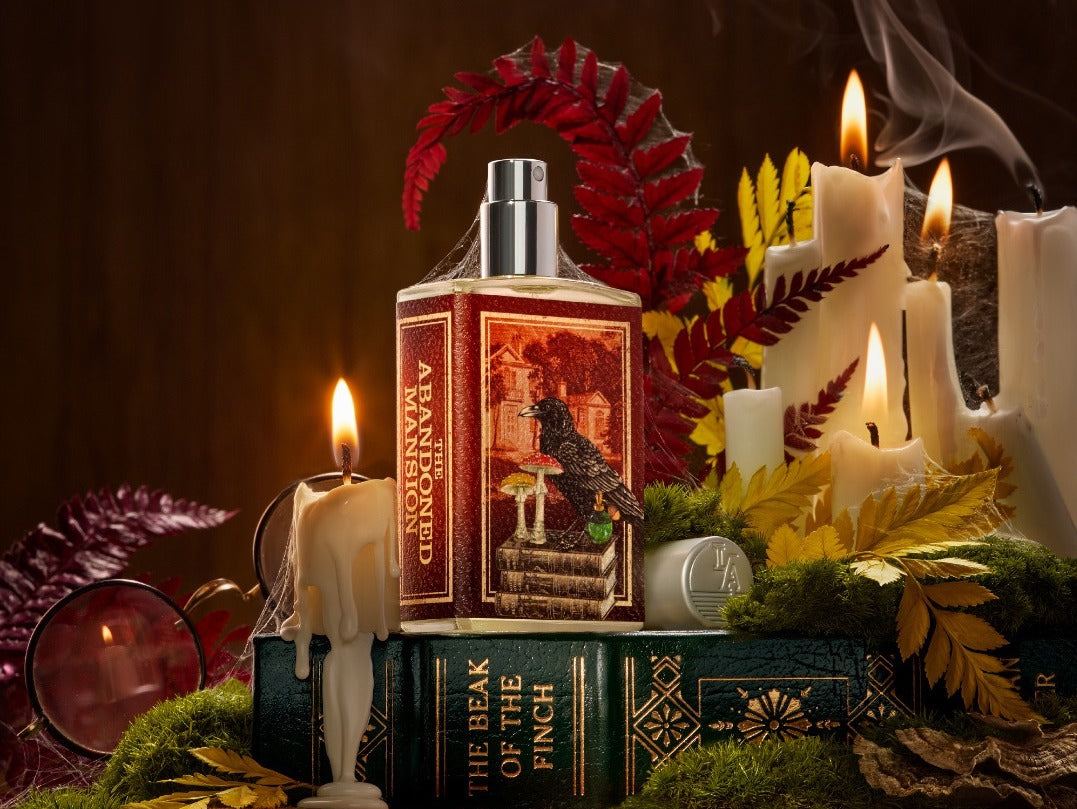 The Abandoned Mansion perfume bottle with a label depicting a raven standing on books with mushrooms. Bottle stands on an old book amidst melting candles, moss and dried ferns.