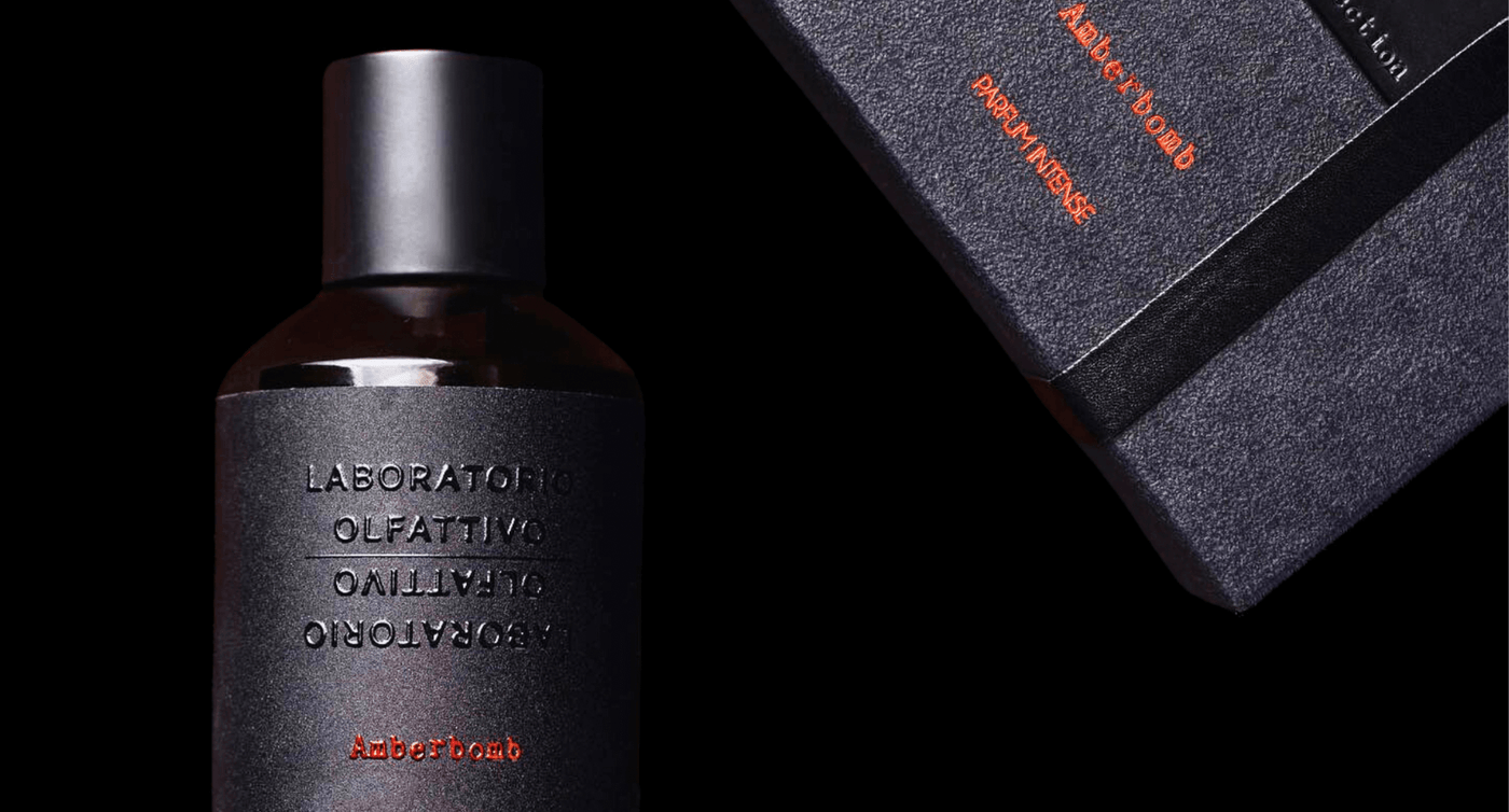 Laboratorio Olfattivo Amberbomb dark glass perfume bottle with black label and red font. Black fragrance box floating off top left corner, black background.