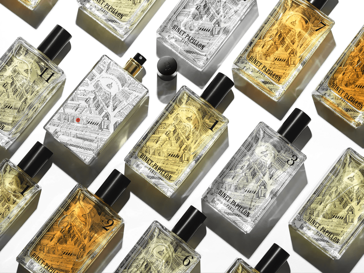 Many Binet-Papillon perfume bottles, placed diagonally on a white surface. Bottles are rectangular clear glass with printed with old drawings of Paris.