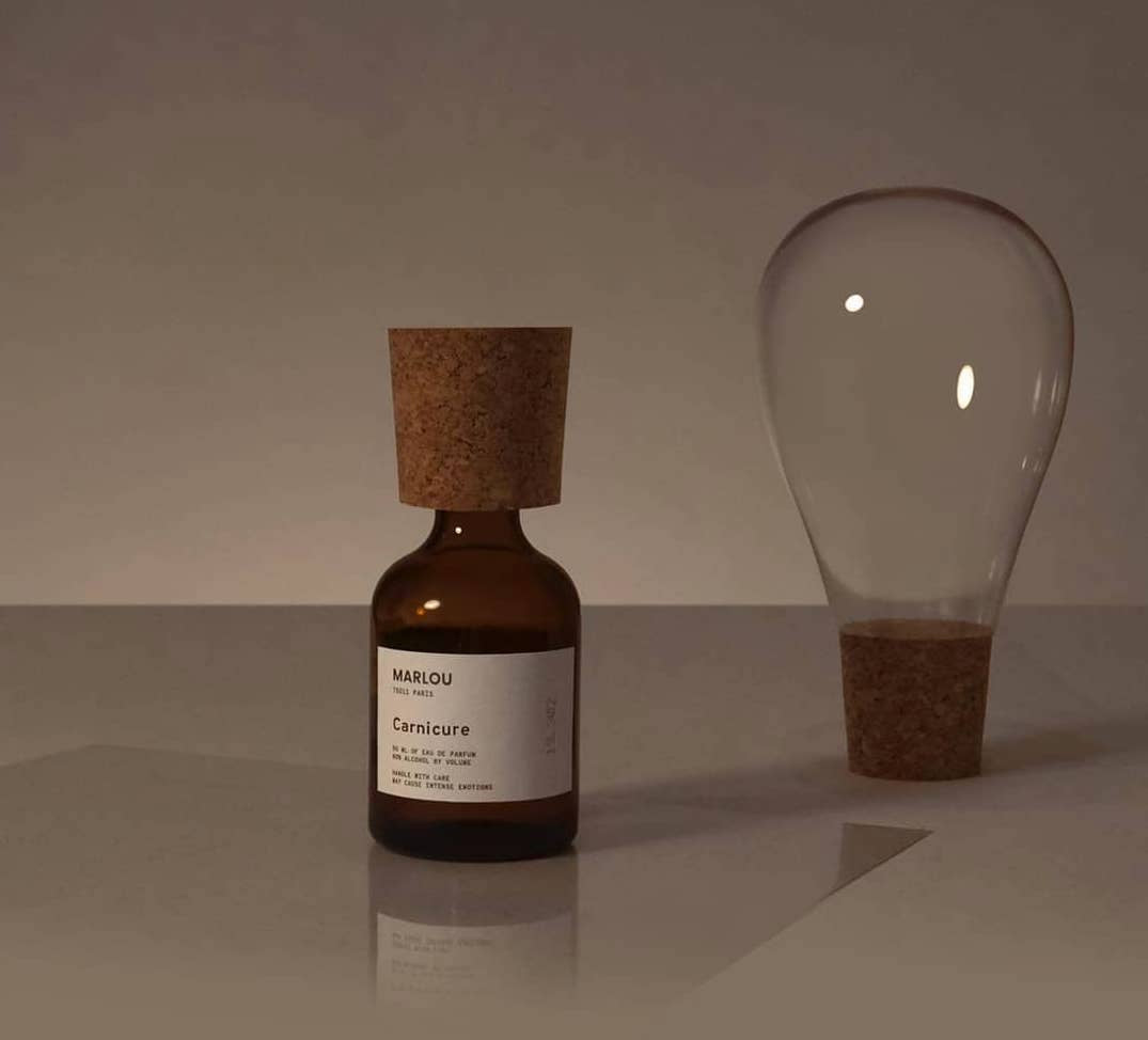 Marlou Carnicure dark amber perfume bottle with modernist cork cap, next to a glass vessel balanced an a second cork cap. Soft taupe-grey background.