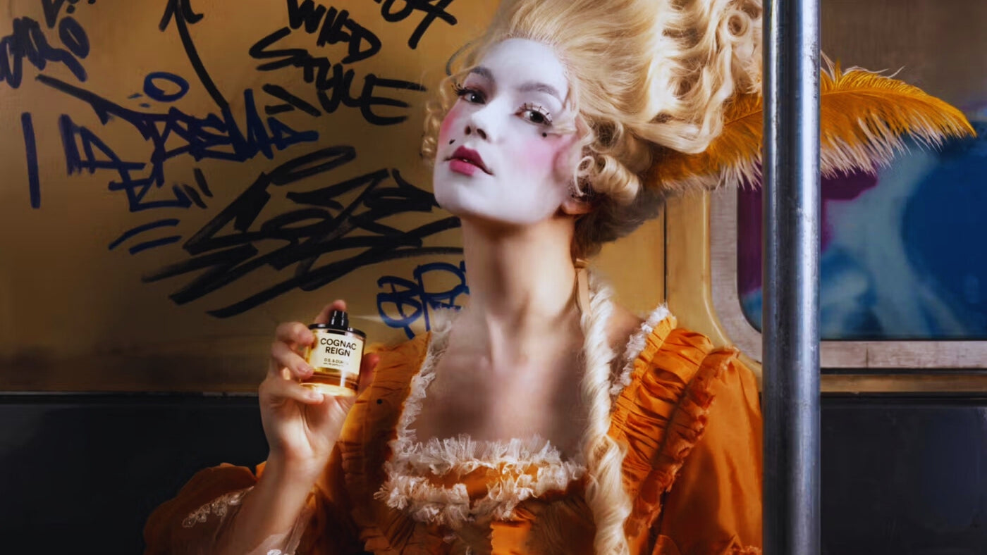 A woman in an orange Baroque gown and tall wig, spraying DS & Durga Cognac Reign, inside a NYC subway car.