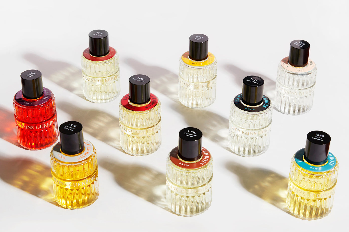 Six Les Bains Guerbois ridged clear glass perfume bottles with black caps on a pale grey surface and background.