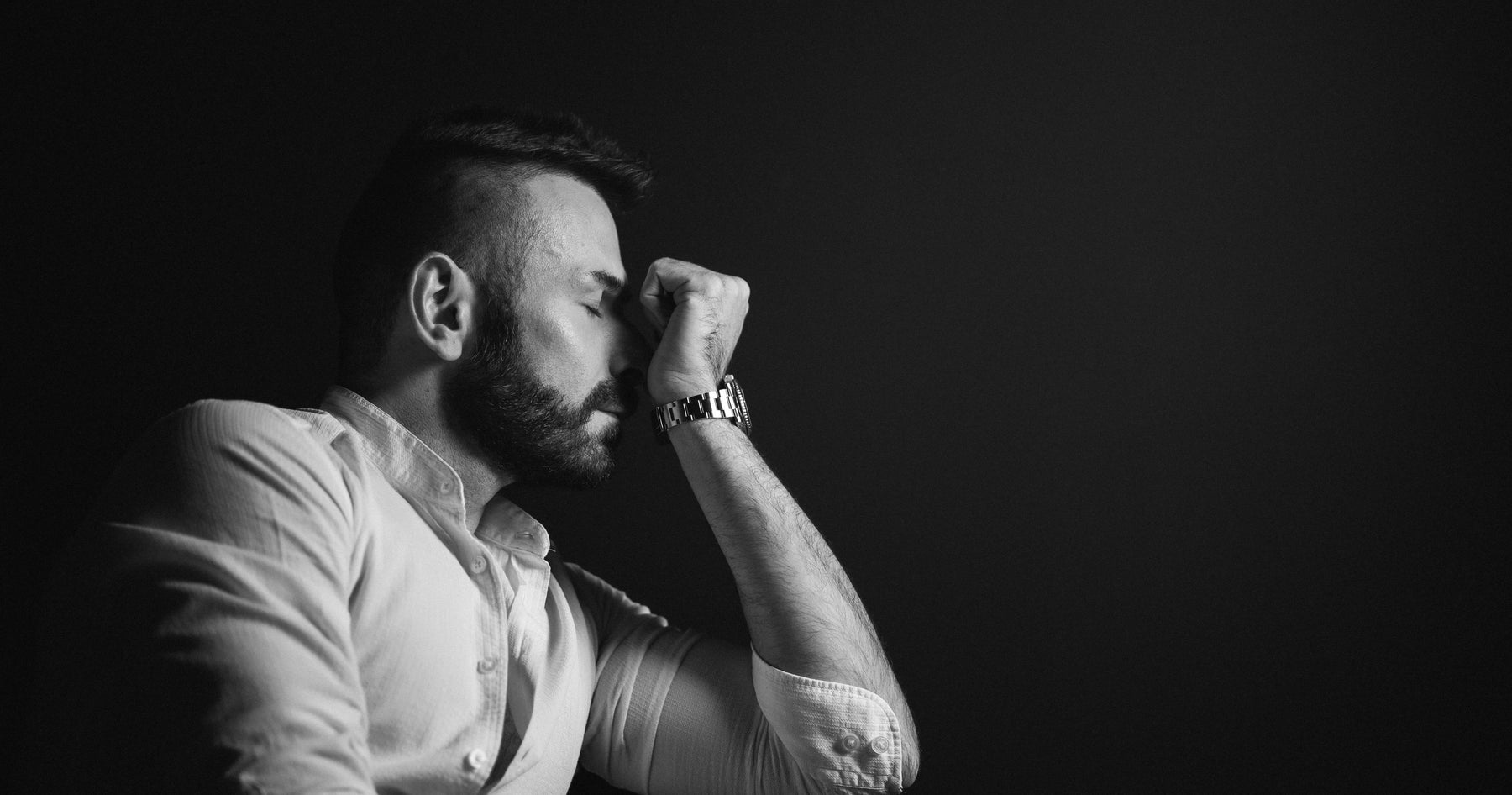 Simone Andreoli in profile, wearing a white collared shirt, smelling his wrist. Black and white with black background.
